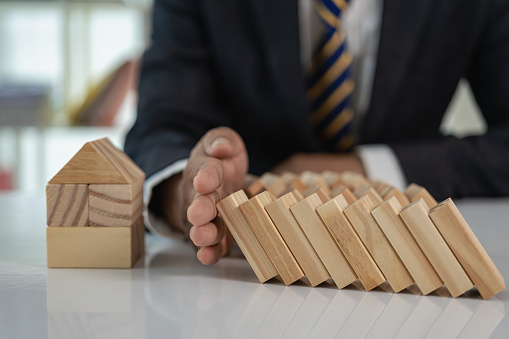 Cropped View Of Risk Manager Protecting House Model From Falling Wooden Blocks With Hands, Home Insurance And Safety. Businessman's Hands Stop Wooden Blocks From Falling Onto The Model House.